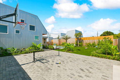 Sunny modern backyard with metal-clad house, paved patio and basketball hoop, portable net and paddles, pool, palm trees and fenced landscaped lawn