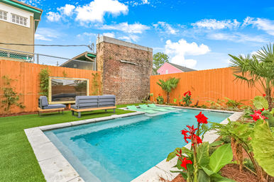 Sunny urban backyard pool with turquoise water, lounge chairs and gray outdoor sofa on artificial turf, orange wooden fence, brick wall, and red tropical flowers
