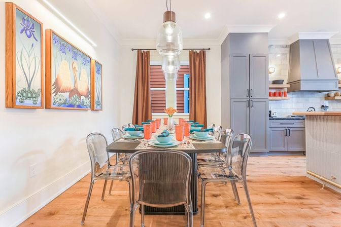 Bright modern open-concept dining room set for eight with clear acrylic chairs, turquoise place settings and coral glasses on a dark wood table, pendant lighting, warm wood floors, gray kitchen cabinets, and colorful bird-and-turtle wall art.