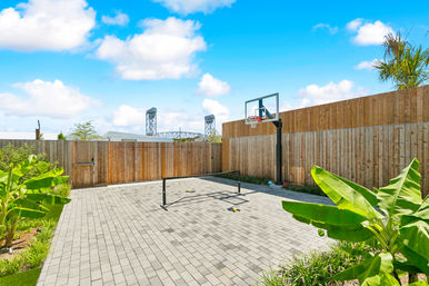 Sunny urban backyard with a paved mini sports court, portable basketball hoop and net, tall wooden fence, tropical banana plants and a bridge silhouette under a bright blue sky.