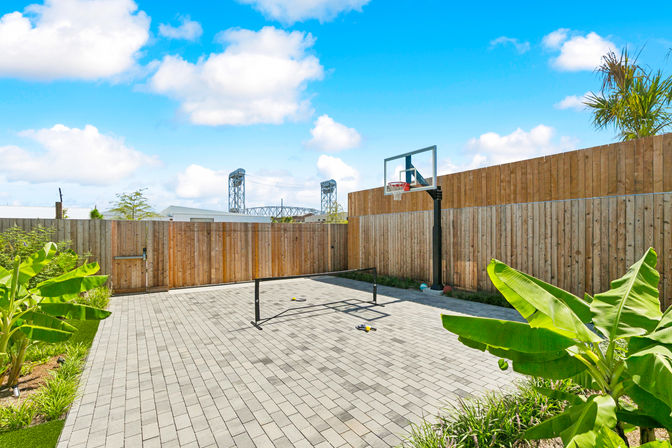 Sunny urban backyard with a paved mini sports court, portable basketball hoop and net, tall wooden fence, tropical banana plants and a bridge silhouette under a bright blue sky.