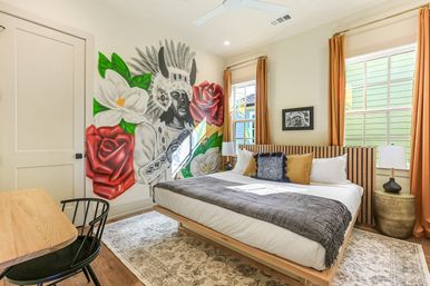 Bright modern bedroom with king bed, wooden slatted headboard, amber curtains, patterned rug and a bold, colorful mural of a headdress-wearing figure surrounded by red roses and white flowers.