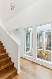 Bright contemporary interior stairwell with warm wooden steps and wide-plank oak floors, crisp white walls and tall windows overlooking a small planted courtyard, flooded with natural light.
