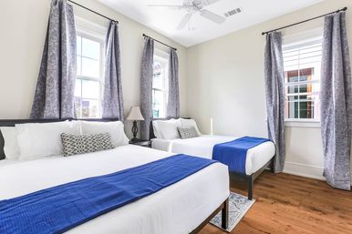 Bright sunny bedroom with two double beds in crisp white linens and blue runners, gray patterned curtains at three windows, hardwood floors, decorative pillows, bedside lamp and ceiling fan.