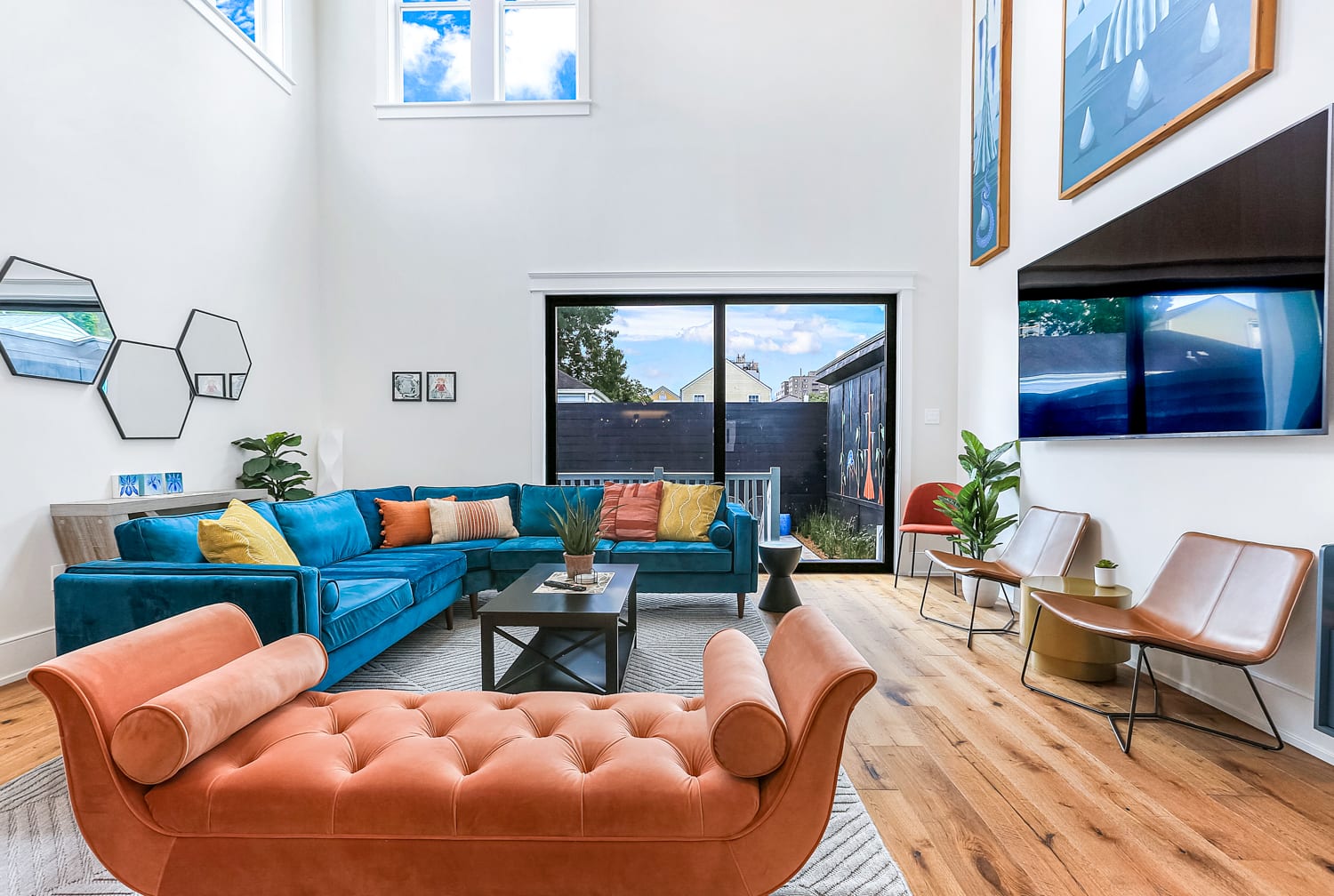 Bright modern two-story living room in an urban townhouse with a teal sectional sofa, coral tufted chaise, tan leather chairs, wood floors, geometric mirrors, plants, coffee table, and a large sliding glass door opening to a small fenced patio.