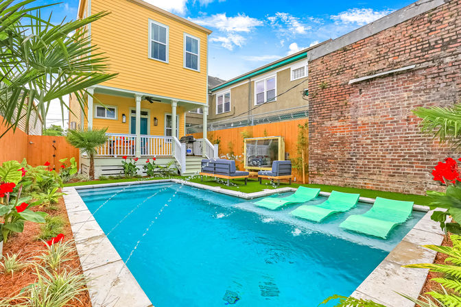 Sunny urban backyard with a turquoise plunge pool, four green in-pool loungers and water jets, outdoor seating on artificial turf, tropical plants, yellow two-story house and exposed brick wall.