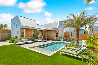 Sunny modern coastal backyard with rectangular swimming pool, palm tree, green chaise lounges on turf, outdoor sofas on a wooden deck, and a wood‑clad house with corrugated metal roof — tropical vacation‑style setting.