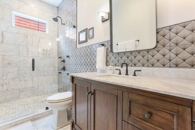 Bright modern bathroom with glass-enclosed marble shower, fan-pattern gray tile backsplash, dark wood vanity with marble countertop and matte black fixtures