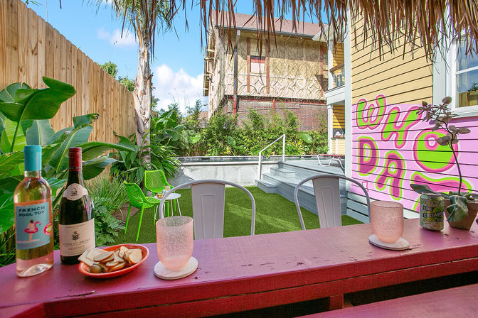 Colorful urban backyard patio with a pink bar holding wine bottles, glasses and snacks, metal chairs facing artificial turf and a raised plunge pool, tropical plants and a yellow house wall with a bright pink-and-green mural.