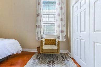Cozy bedroom corner with mustard velvet tufted armchair beneath a sunny window, round-patterned curtains, white closet doors, hardwood floor and leaf-print rug.