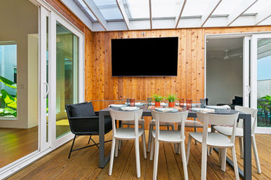 Cozy modern covered patio dining area on a wooden deck with a wood-paneled wall and wall-mounted TV, set table with white chairs and potted plants, framed by sliding glass doors to the interior.