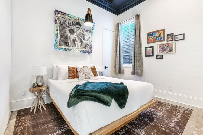 Bright, cozy modern bedroom with white walls and a navy ceiling, low wooden platform bed dressed in white linens, patterned pillows and a folded green velvet throw, gallery wall art, pendant light and a small wooden side table with lamp and succulent.