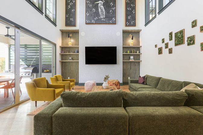 Sunlit modern double-height living room with a green sectional sofa, two mustard-yellow armchairs, wall-mounted TV flanked by wood-slatted shelves, large heron artwork above, and sliding glass doors opening to a covered patio with a grill.
