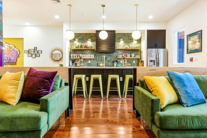 Bright modern open-plan kitchen and living room with green velvet sofas, colorful throw pillows, island with four lime bar stools, green subway tile backsplash, globe pendant lights and warm hardwood floors