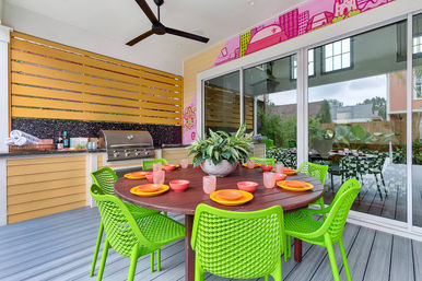 Covered backyard patio dining area with round wooden table set for eight, vibrant lime-green chairs, colorful plates and cups, potted centerpiece, built-in stainless-steel grill and sliding glass doors to a garden.