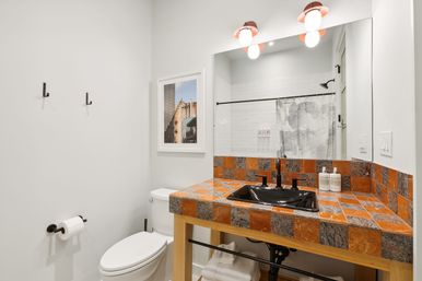 Bright modern apartment bathroom with orange-and-gray checkerboard tile vanity, black sink and faucet, large mirror with exposed-bulb lights, white toilet, framed city photo and shower reflected in the mirror.