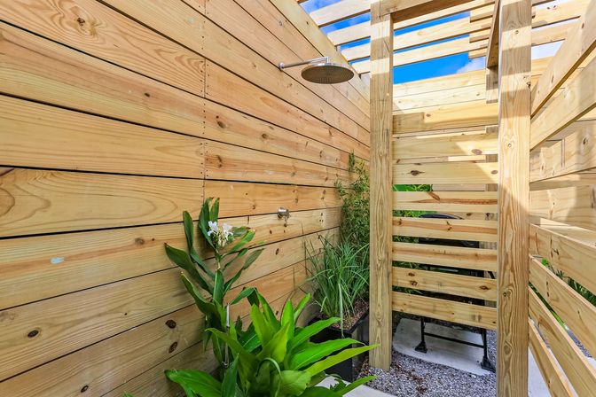 Sunlit backyard outdoor shower with overhead rain showerhead, light wooden slat privacy walls and open slatted roof, lush potted tropical plants, and gravel floor.