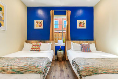 Cozy modern twin bedroom in an urban apartment with a bold blue accent wall, two white beds with wooden slat headboards, patterned pillows and throws, mustard curtains framing a window, bedside lamp on a decorative table, and hardwood floors.