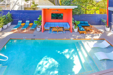 Bright resort-style outdoor swimming pool with coral cabana, blue cushioned lounge seating, sun loungers and in-pool loungers on a sunny tree-shaded deck with potted plants