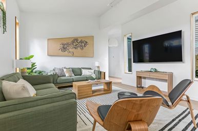 Bright, airy modern living room with green mid-century sofas, wooden coffee table and curved wood accent chair, wall-mounted flat-screen TV, striped rug, indoor plants and minimalist wood decor.