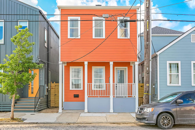 Bright orange two-story row house with white trim and a small front porch between gray and blue homes on an urban street, parked van and overhead utility wires visible.
