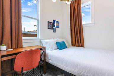 Cozy bright bedroom with crisp white bed and teal accent pillow, wooden desk with orange chair and small succulent beside two windows framed by brown curtains, simple wall art and an urban rooftop view.