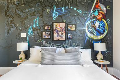 Boutique hotel bedroom with a white bed and patterned lumbar pillow against a dark mural wall depicting a colorful tuba player, neon musical notes, sketched city skyline and framed street photos, flanked by matching bedside lamps on round tables.