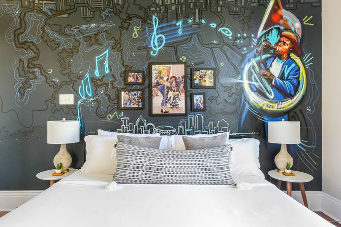 Boutique hotel bedroom with a white bed and patterned lumbar pillow against a dark mural wall depicting a colorful tuba player, neon musical notes, sketched city skyline and framed street photos, flanked by matching bedside lamps on round tables.