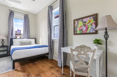Cheerful sunlit urban bedroom with hardwood floors, white bed with blue runner, gray patterned curtains, city-view window, white writing desk with ornate chair, floor lamp and framed colorful artwork