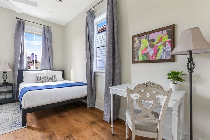 Cheerful sunlit urban bedroom with hardwood floors, white bed with blue runner, gray patterned curtains, city-view window, white writing desk with ornate chair, floor lamp and framed colorful artwork