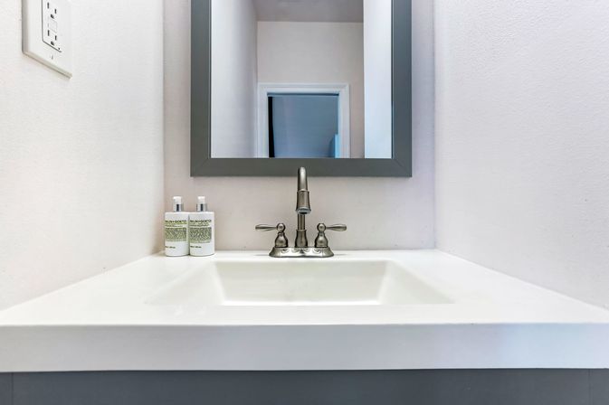 Minimalist white bathroom sink with stainless faucet, gray-framed mirror, and two soap dispensers on a white vanity in a modern powder room