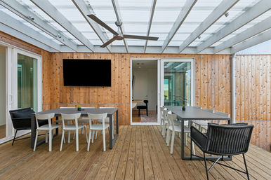 Covered wooden-deck patio with two gray dining tables, white chairs and wicker armchairs, wood-paneled walls, wall-mounted TV, ceiling fan and sliding glass doors.