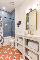 Bright modern bathroom with blue subway-tiled tub-shower, brass fixtures, white open-shelf vanity with folded towels, and bold orange geometric floor tiles.