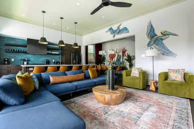 Open-concept modern living room and kitchen with deep blue sectional, mustard barstools, teal tile backsplash, olive velvet armchairs, round wooden coffee table, patterned rug and playful pelican wall murals