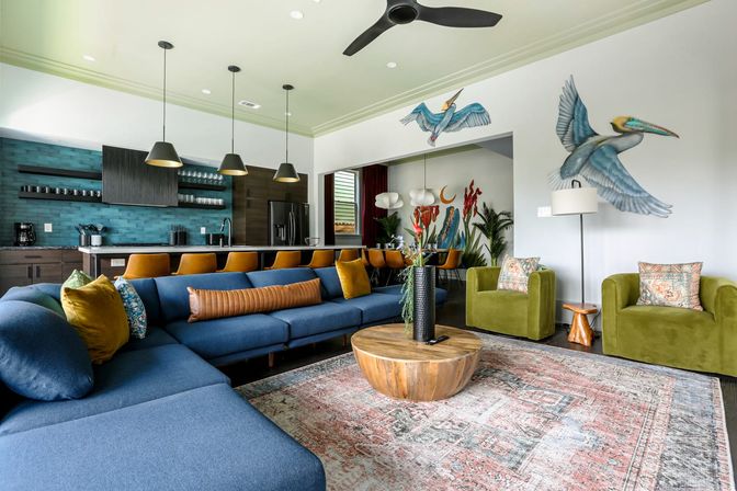 Open-concept modern living room and kitchen with deep blue sectional, mustard barstools, teal tile backsplash, olive velvet armchairs, round wooden coffee table, patterned rug and playful pelican wall murals