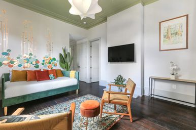 Sunlit modern living room with green velvet daybed and colorful throw pillows, whimsical floral wall mural, mid-century wooden chairs, orange velvet ottoman on a patterned rug, wall-mounted TV, hardwood floors and potted plants.