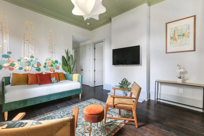Sunlit modern living room with green velvet daybed and colorful throw pillows, whimsical floral wall mural, mid-century wooden chairs, orange velvet ottoman on a patterned rug, wall-mounted TV, hardwood floors and potted plants.