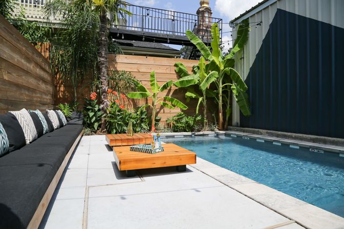 Urban backyard plunge pool beside a modern patio with a black cushioned bench and patterned pillows, low wooden coffee tables, banana plants, palm, and string lights.