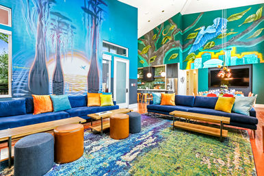 Vibrant modern community lounge with high ceilings and colorful ocean-and-tree murals, two long navy sofas with bright throw pillows, wooden benches, leather and fabric ottomans on a multicolored area rug, and an open kitchen with bar stools and pendant lights.