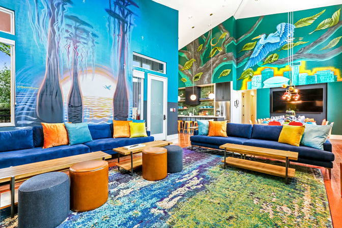 Vibrant modern community lounge with high ceilings and colorful ocean-and-tree murals, two long navy sofas with bright throw pillows, wooden benches, leather and fabric ottomans on a multicolored area rug, and an open kitchen with bar stools and pendant lights.