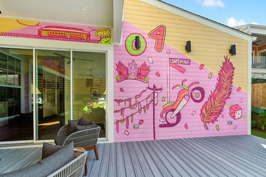 Outdoor patio with sliding glass door and a bright pink Louisiana-themed mural featuring a feather, beads, dice, motorcycle and train on a yellow house exterior.