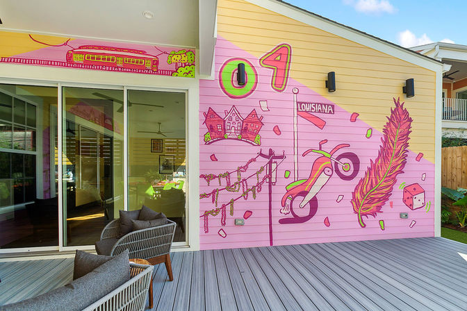 Outdoor patio with sliding glass door and a bright pink Louisiana-themed mural featuring a feather, beads, dice, motorcycle and train on a yellow house exterior.
