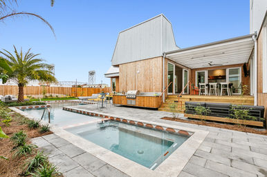 Modern wood-clad home with covered deck and outdoor dining, built-in grill, rectangular pool and spa, palm tree, and bridge visible in the background.