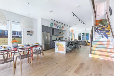 Bright, cheerful open-concept kitchen and dining room with hardwood floors, long island with barstools, stainless refrigerator, red dining table, pendant lights, and a colorful mural-painted staircase.