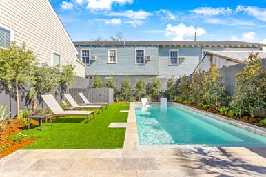 Sunny urban backyard with a long rectangular swimming pool, stone patio and artificial turf with three lounge chairs, gray privacy fence, landscaping and a two-story light-blue house behind.