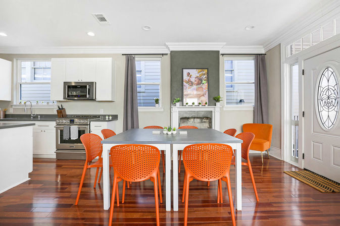 Bright open-plan kitchen-dining room with polished hardwood floors, white cabinetry and stainless range, gray dining table surrounded by vibrant orange modern chairs, decorative fireplace with artwork, and glass-paneled entry door.