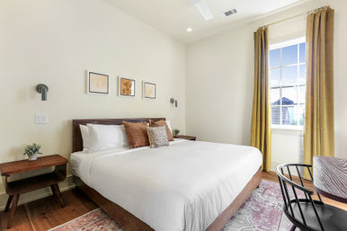 Sunlit modern bedroom with a wooden king bed dressed in crisp white linens, mid-century nightstands, mustard curtains, hardwood floors and framed wall art.