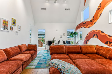 Sunlit open-plan modern living room with a large rust-orange velvet sectional, blue-green patterned rug, vaulted white ceiling, retro orange wave wall mural, dining table, windows and potted plants.