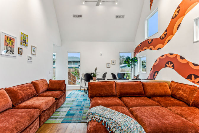Sunlit open-plan modern living room with a large rust-orange velvet sectional, blue-green patterned rug, vaulted white ceiling, retro orange wave wall mural, dining table, windows and potted plants.