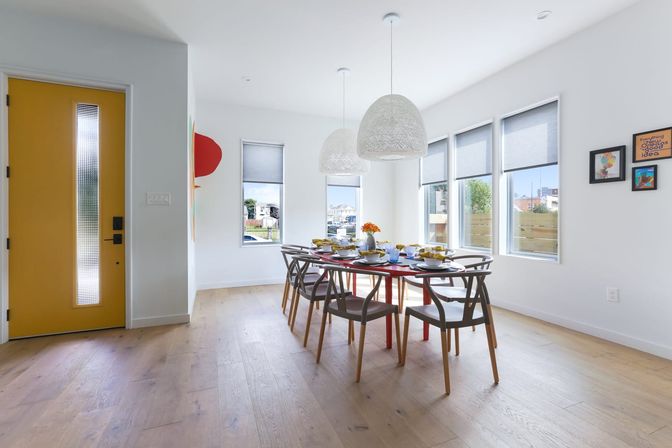 Bright modern dining room in a sunny urban home with a yellow front door, large windows with roller shades, a long red table set for eight, woven pendant lights and light hardwood floors.